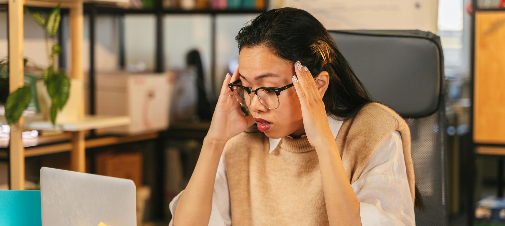 A woman with glasses shows stress while working at a laptop in an office environment.